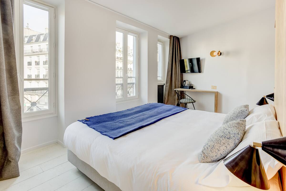 A hotel room with a bed, a TV, a desk, and a view of buildings through the windows.