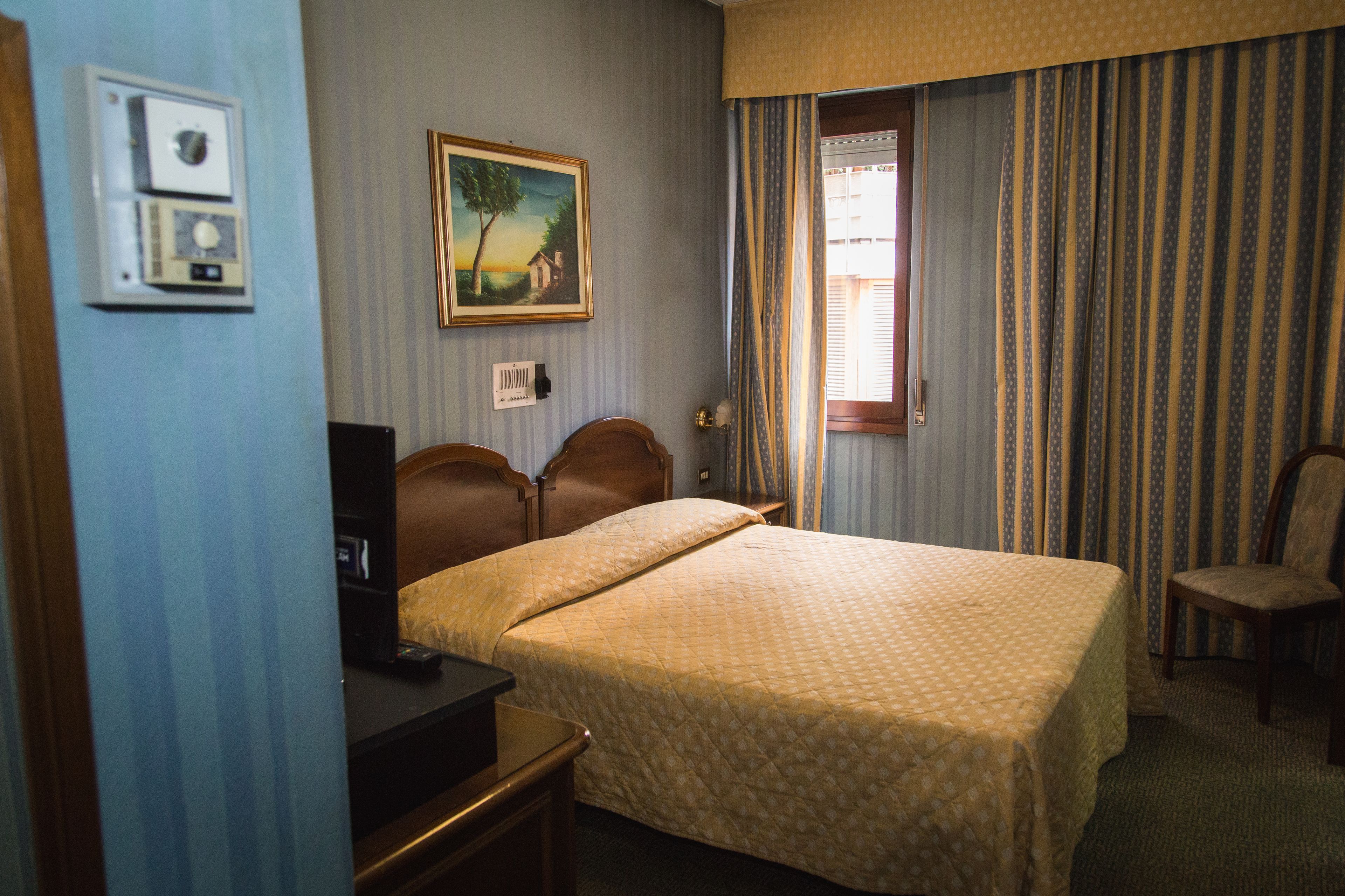 A hotel room with a bed, a desk, a chair, a painting on the wall, and a window with curtains.