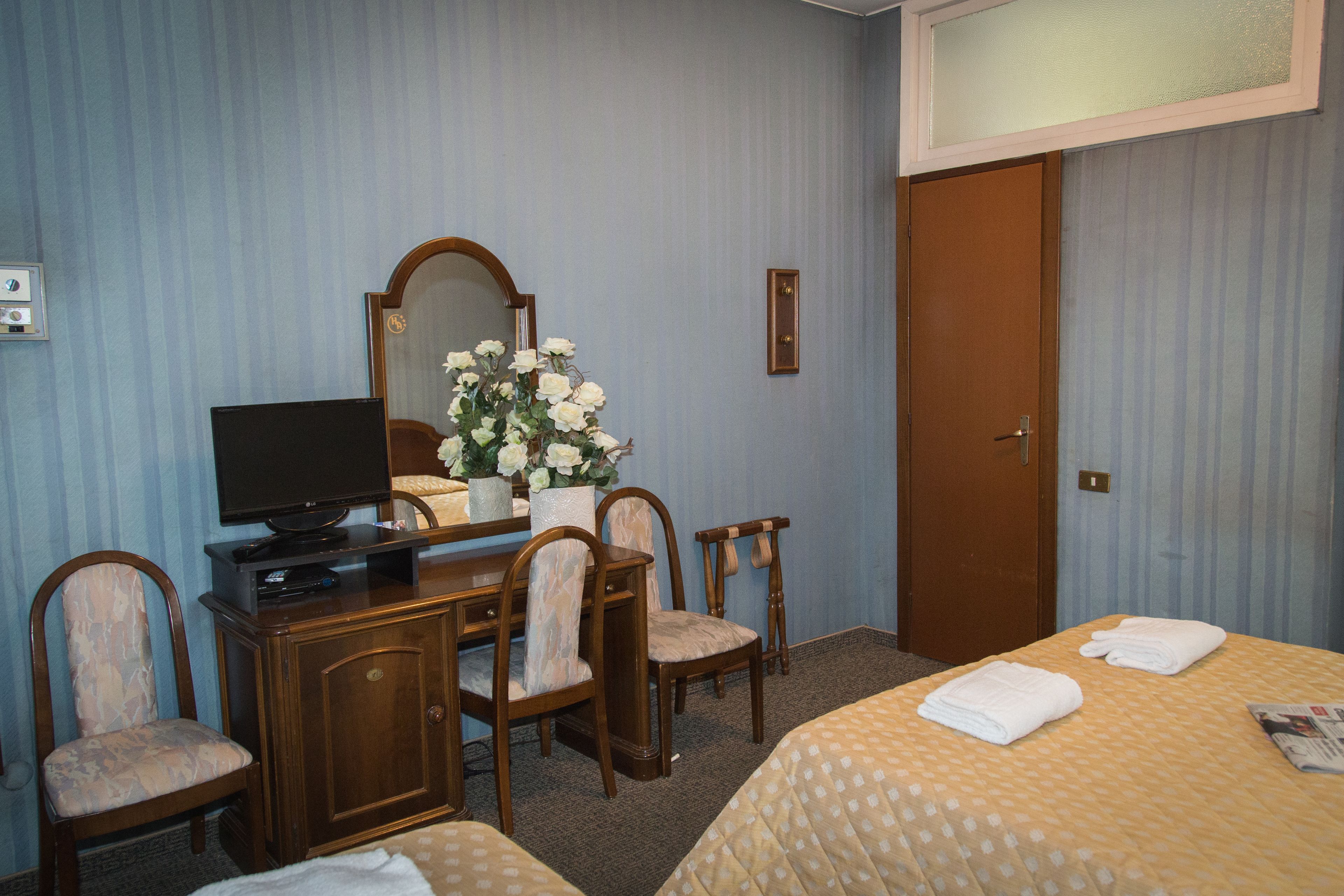 A hotel room with a bed, a desk with a TV, two chairs, a mirror, and a door.