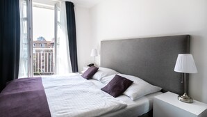 Un cuarto de hotel con una cama grande, una mesita de noche con lámpara y vista a edificios a través de una ventana.