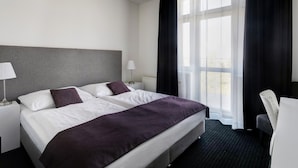 Habitación de hotel con una cama grande, dos mesitas de noche, una lámpara y vistas al exterior a través de una ventana con cortinas.