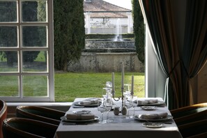 Mesa de comedor dispuesta para seis personas, con velas, platos y copas, con vistas a un jardín con fuente.