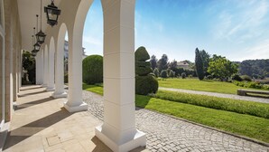 Un paseo cubierto con columnas blancas y faroles colgantes, que conduce a un jardín cuidado con setos y un cielo azul despejado.