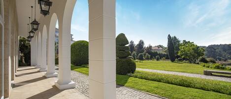 A covered walkway with white columns and hanging lanterns, leading to a well-manicured garden with hedges and a clear blue sky.