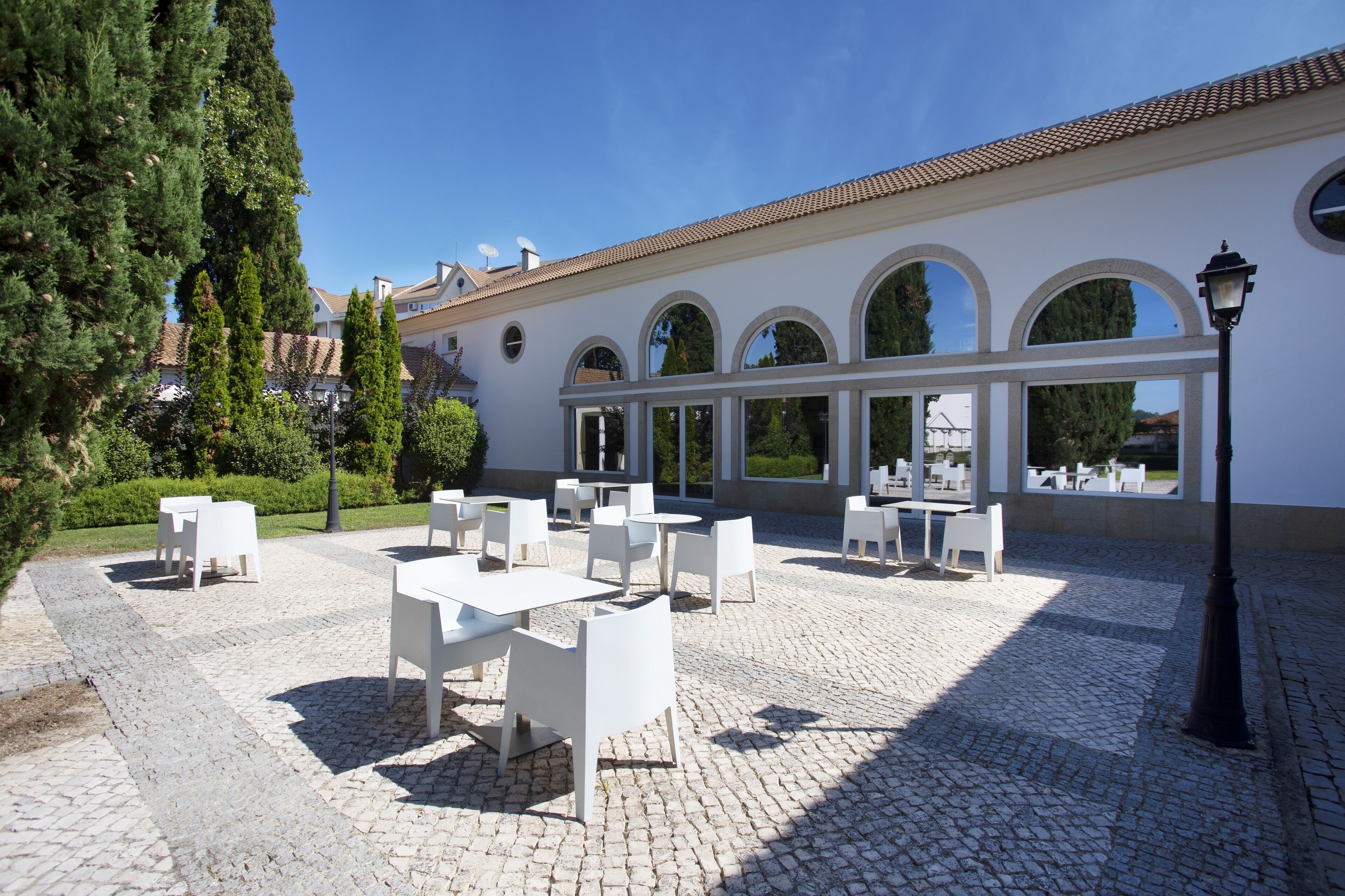 Un patio exterior con mesas y sillas blancas, piso de adoquines y un edificio blanco con ventanas de arco y techo de tejas.