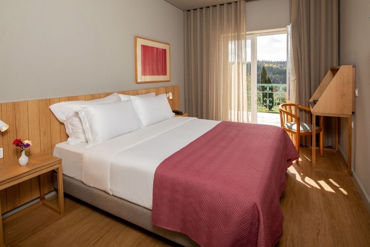 A modern hotel room with a large bed, a desk, and a view of the outdoors.