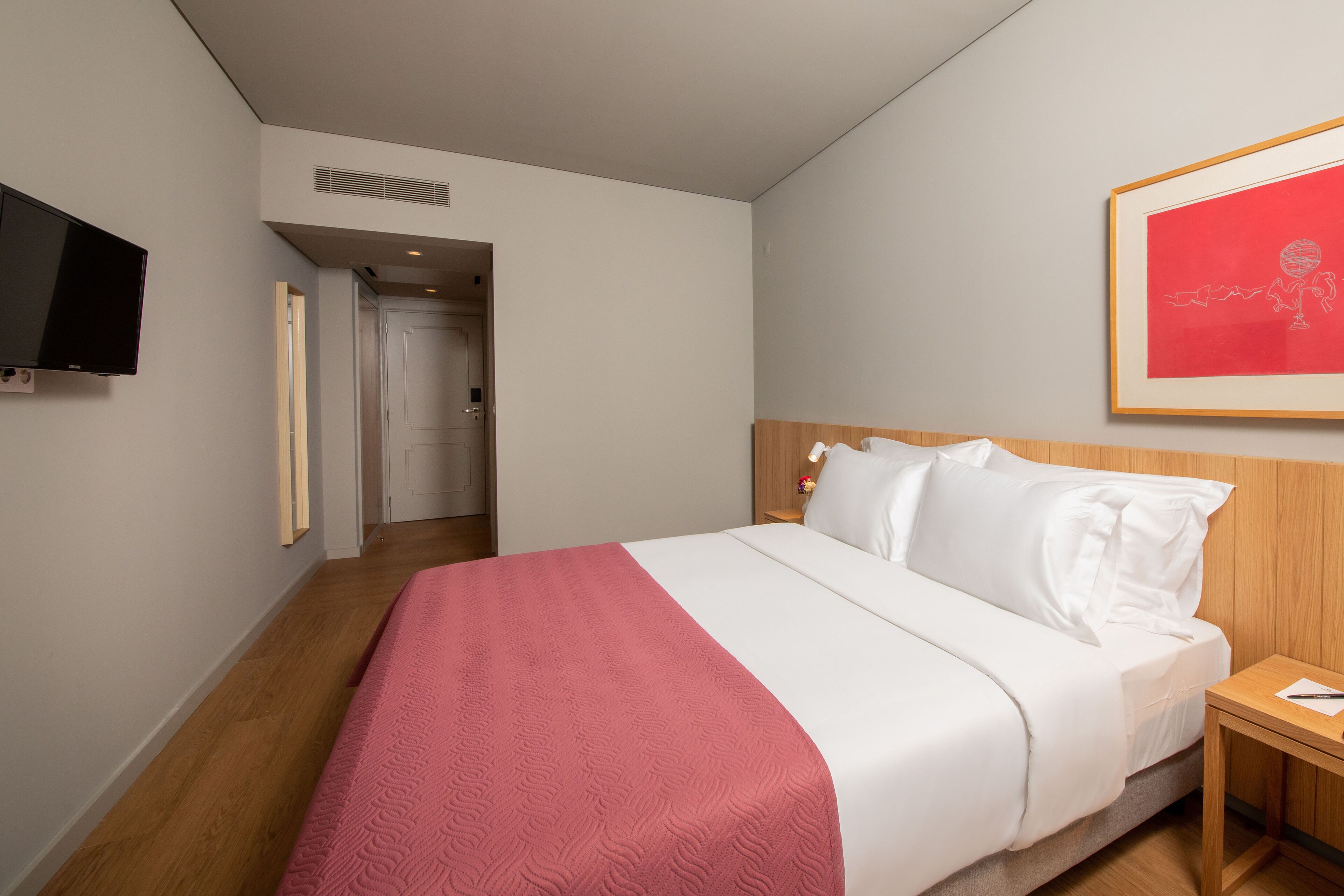 A hotel room with a bed, a TV mounted on the wall, a framed picture, and a wooden nightstand.