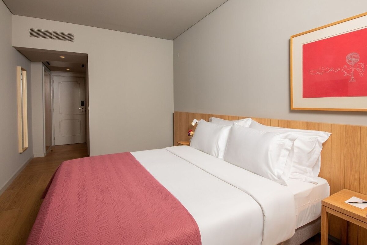 A hotel room with a bed, a nightstand, a framed picture on the wall, and a door leading to another room.