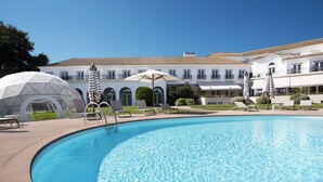 Una piscina con sillas de descanso y sombrillas, rodeada por un edificio con varios balcones y un cielo despejado.