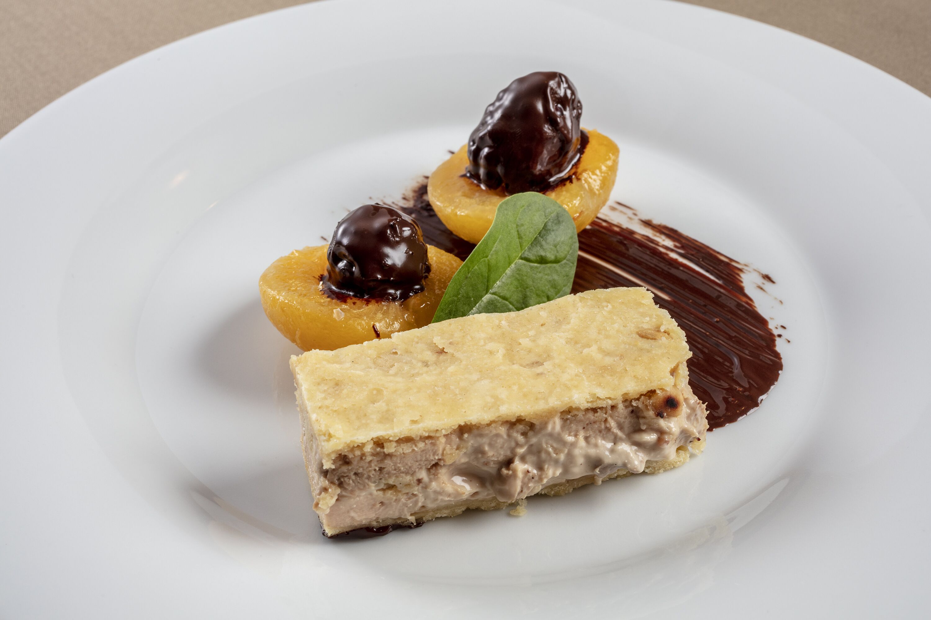 Un plato de postre con una porción de tarta, dos frutas cubiertas de chocolate y una decoración de hoja.