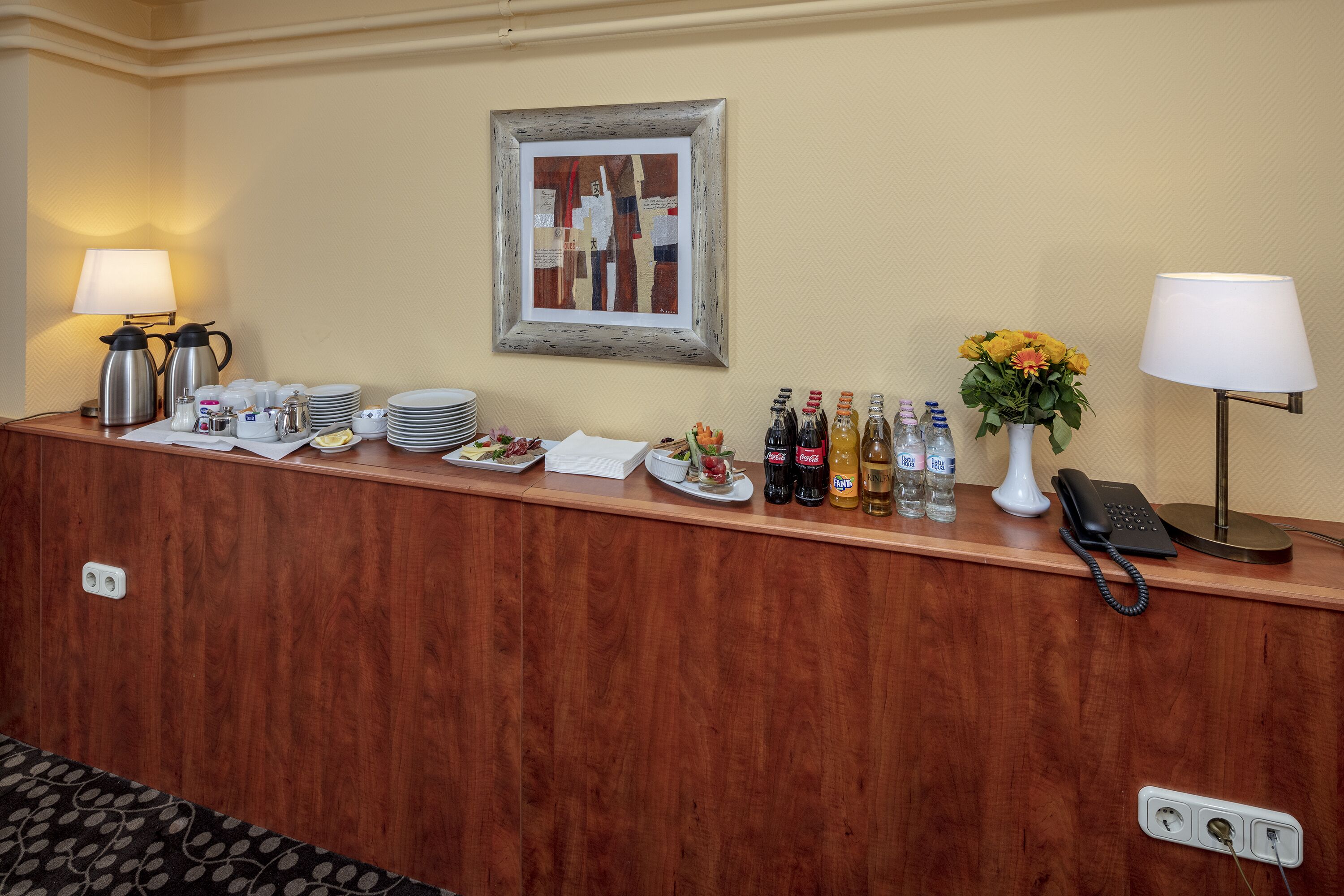 Ein Hotel-Rezeptionsbereich mit einem Holzbüro, einem gerahmten Kunstwerk, einem Telefon und verschiedenen Annehmlichkeiten wie Kaffeeautomaten, Tellern und abgefüllten Getränken.