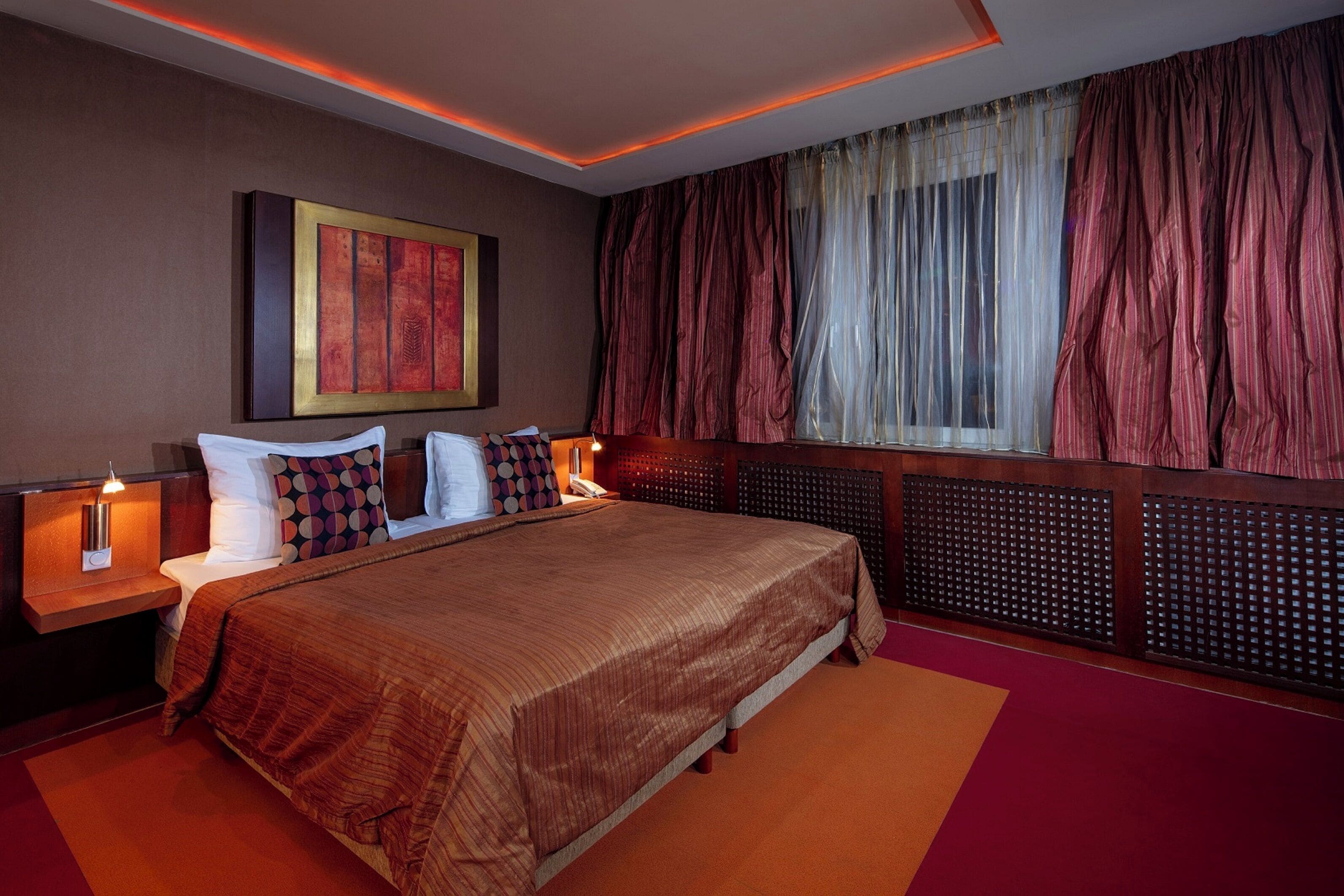 A hotel room with a large bed, a nightstand with a lamp, a wall-mounted artwork, and a window with curtains.
