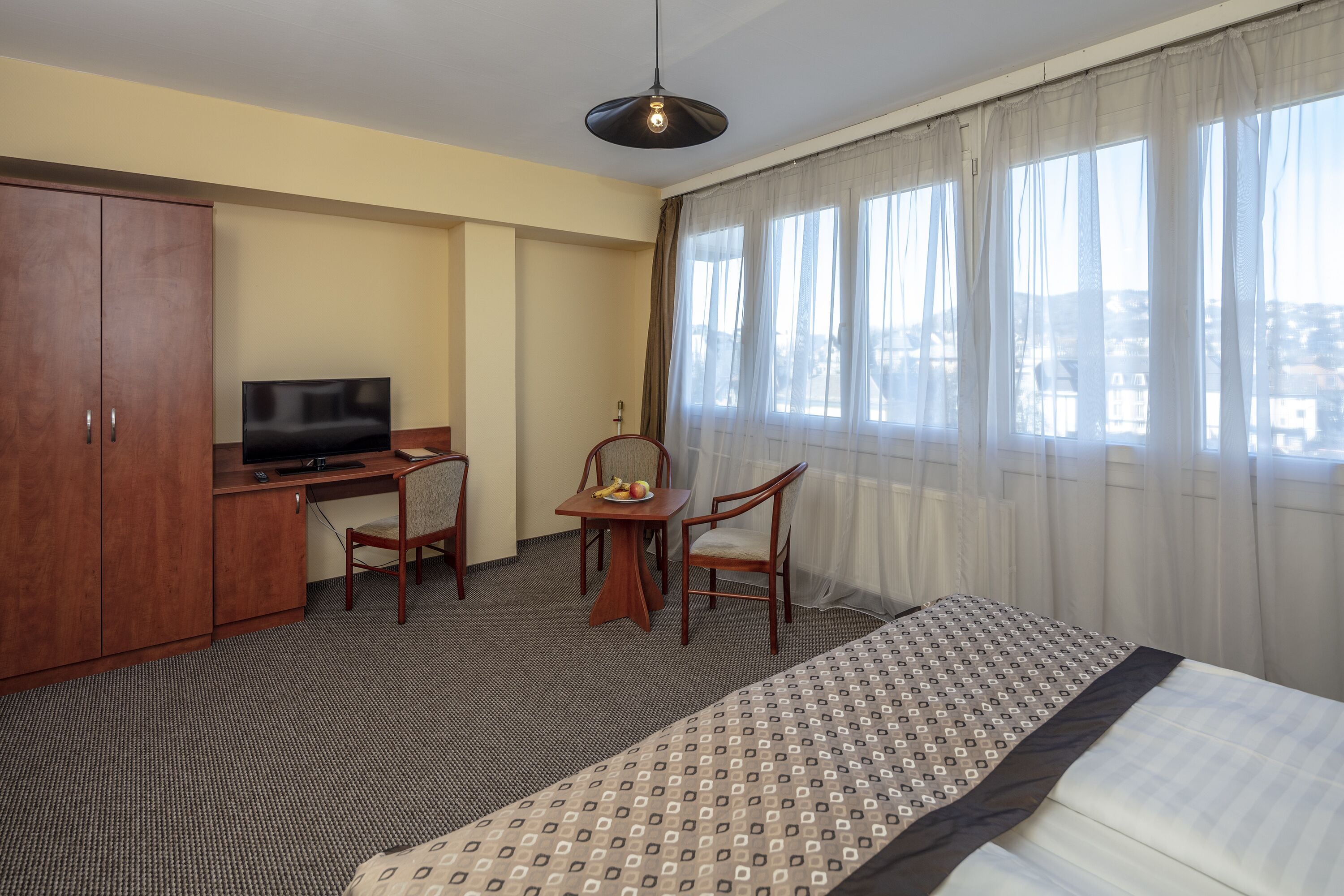 A hotel room with a bed, a desk with a television, a chair, a small table with fruits, and a view of the city through large windows.