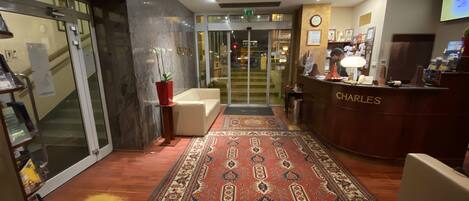 Un hall de hotel con una recepción identificada como 'CHARLES', una alfombra estampada y una escalera que sube a un piso superior.