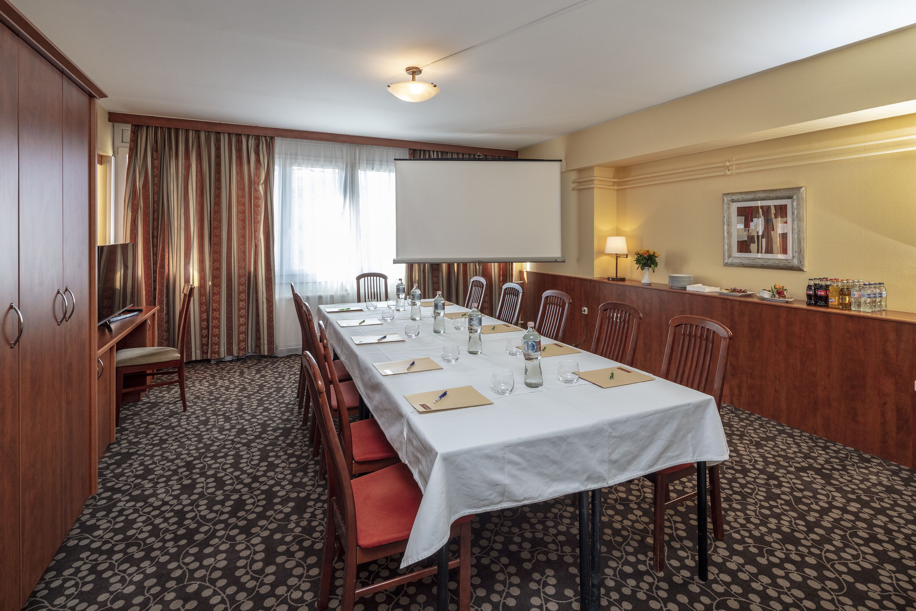 A conference room with a long table, chairs, a projector screen, and a reception area with a counter and refreshments.