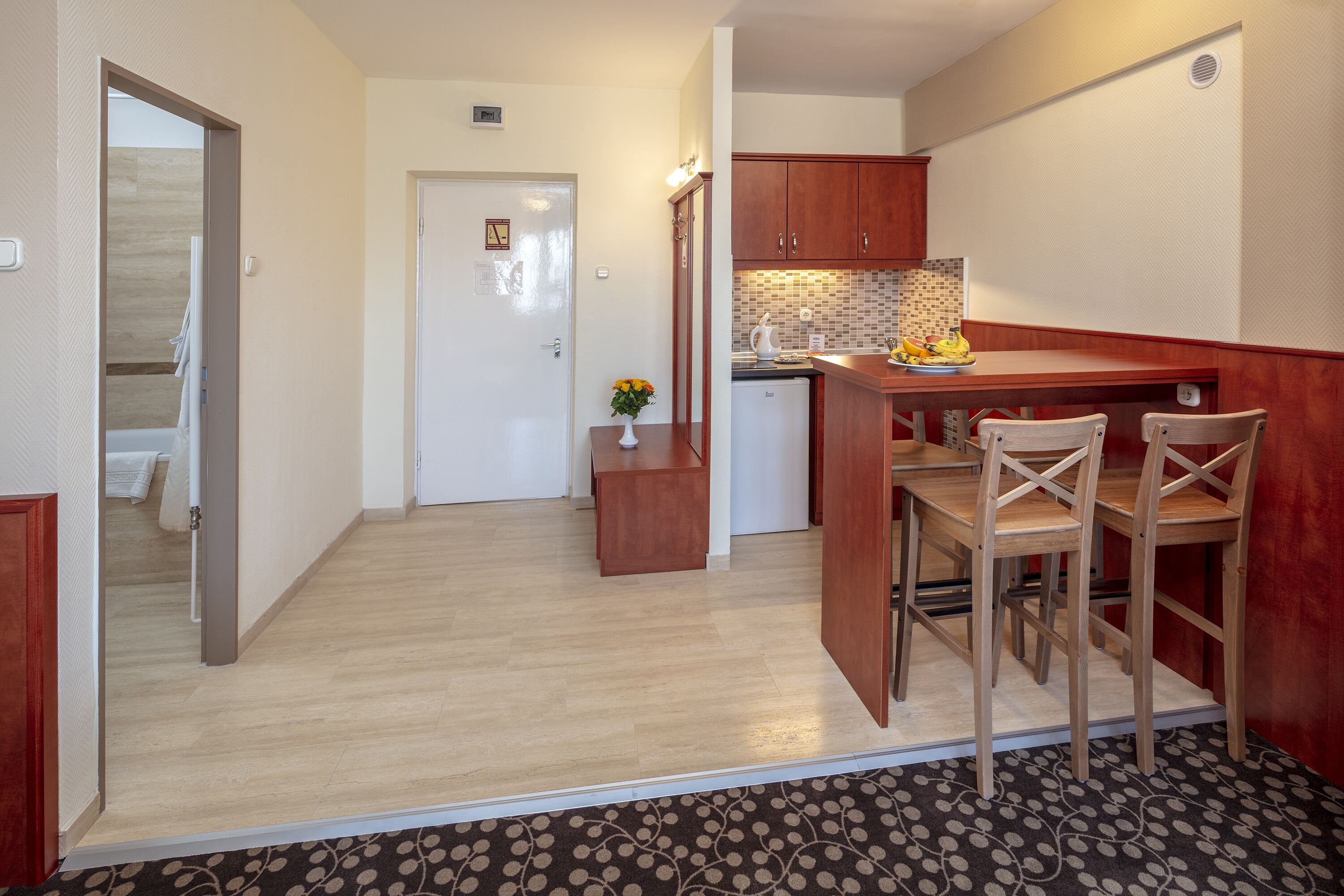 Cocina compacta con armarios de madera, una pequeña mesa de comedor y sillas. Hay un refrigerador, un lavamanos y una encimera con salpicadero. Un jarrón con flores está sobre la encimera.