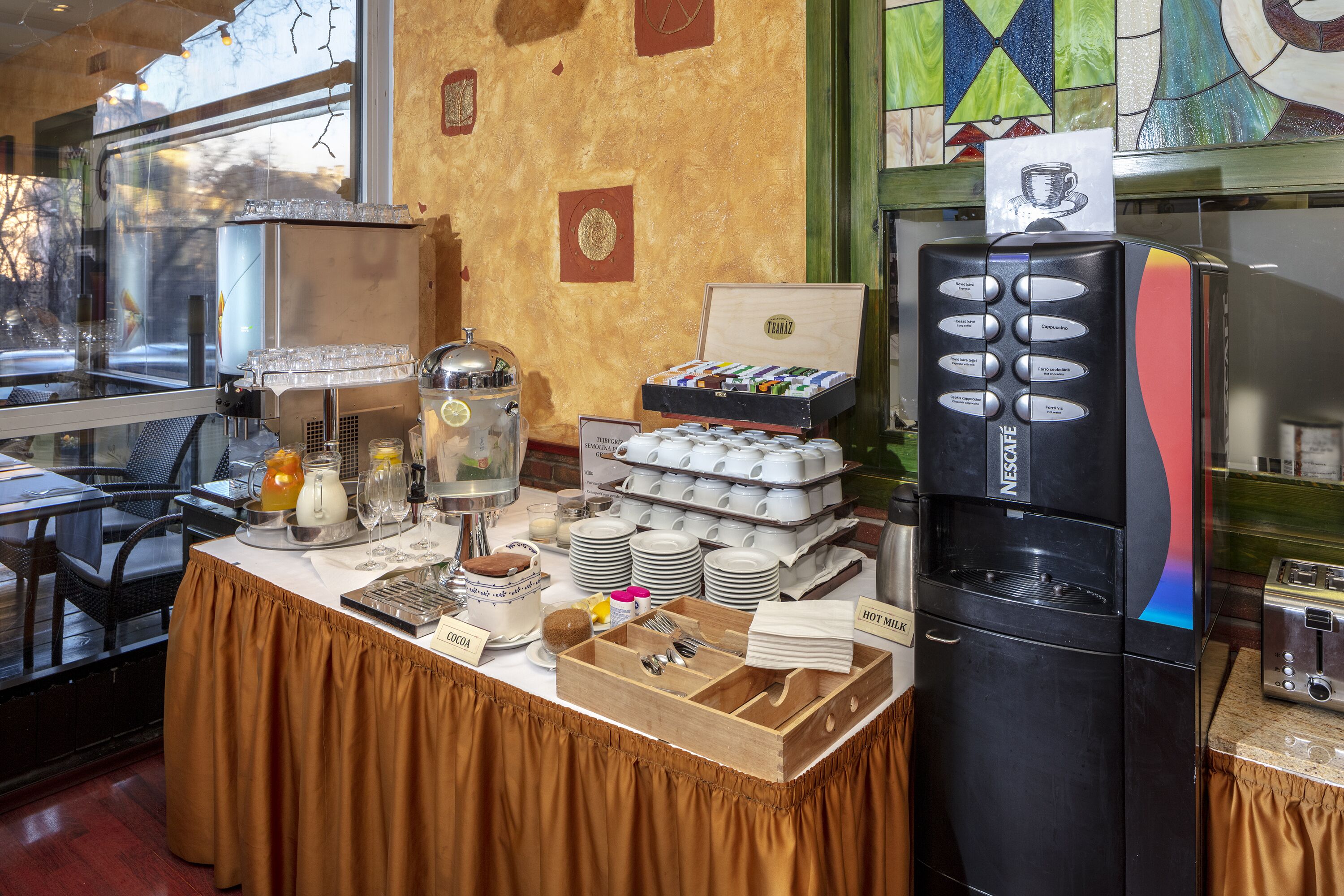 Buffet de café da manhã com máquina de café, espaço para chás e diversas opções de alimentos.