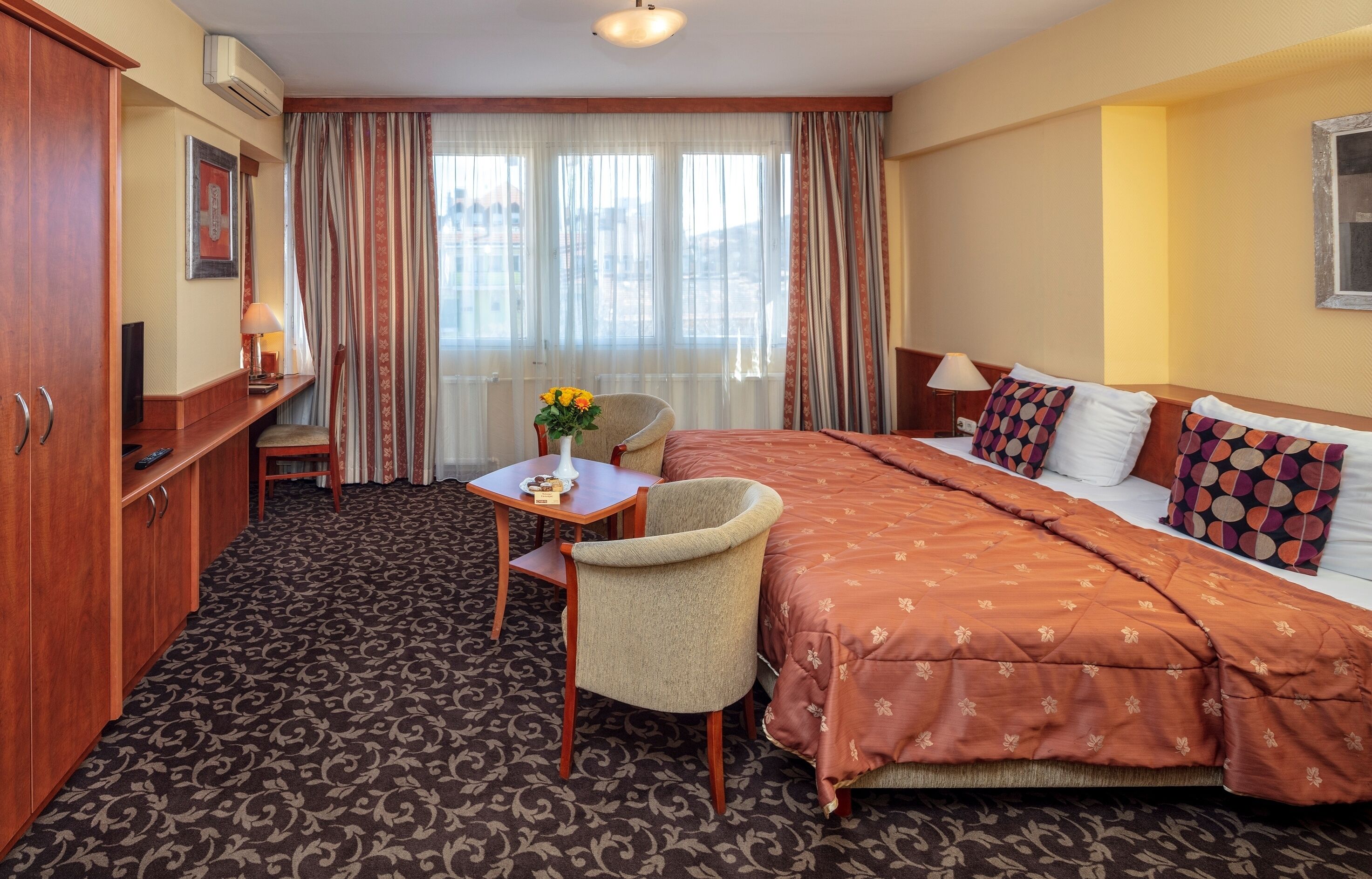 A hotel room with a large bed, a desk with a chair, a small table with a vase of flowers, and a wardrobe.