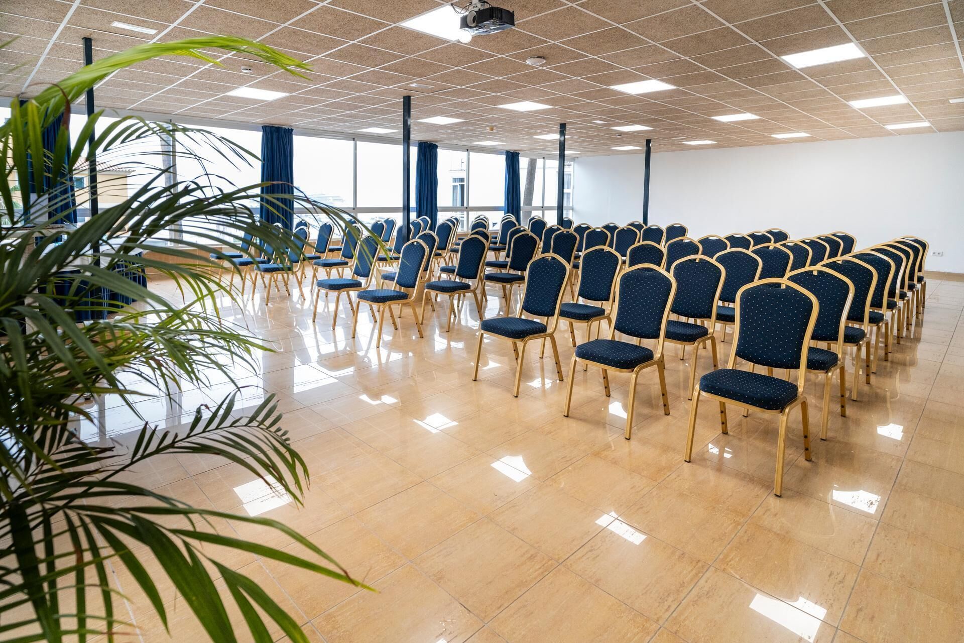 Un salón de conferencias con filas de sillas, un proyector y ventanas amplias.