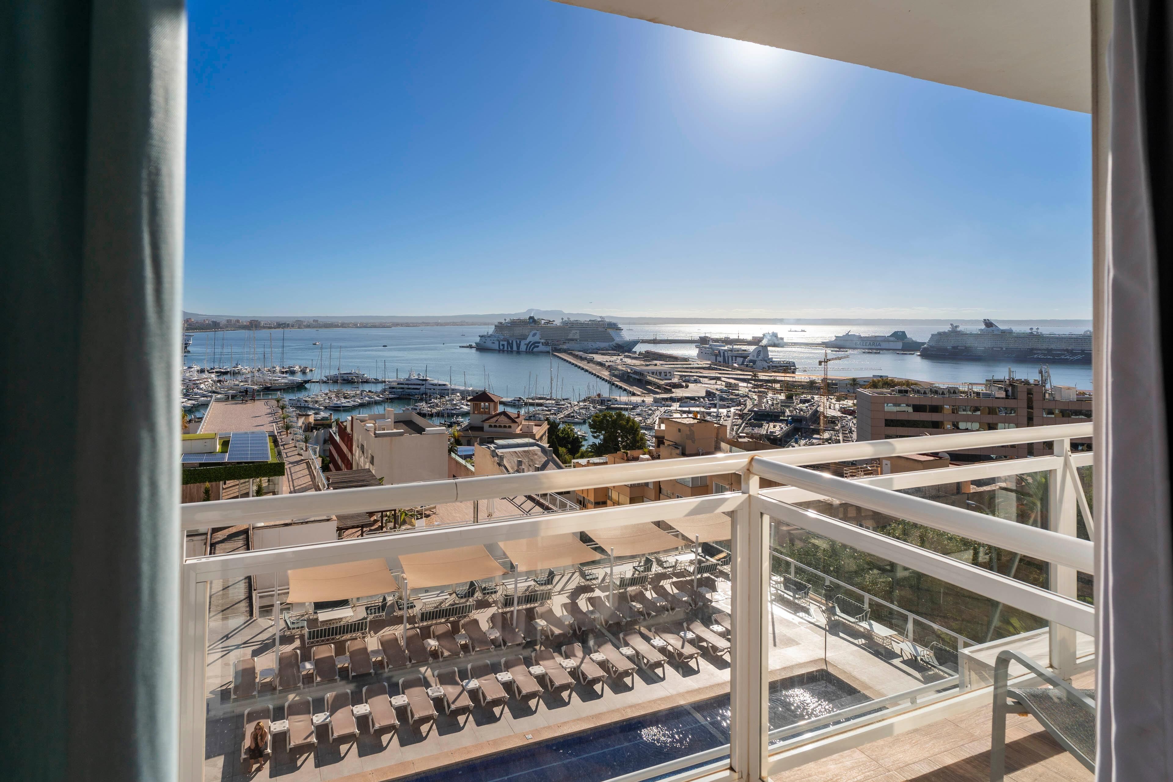 Un balcon avec vue sur un port de plaisance et des bateaux, une piscine avec des chaises longues et un ciel d’un bleu éclatant.