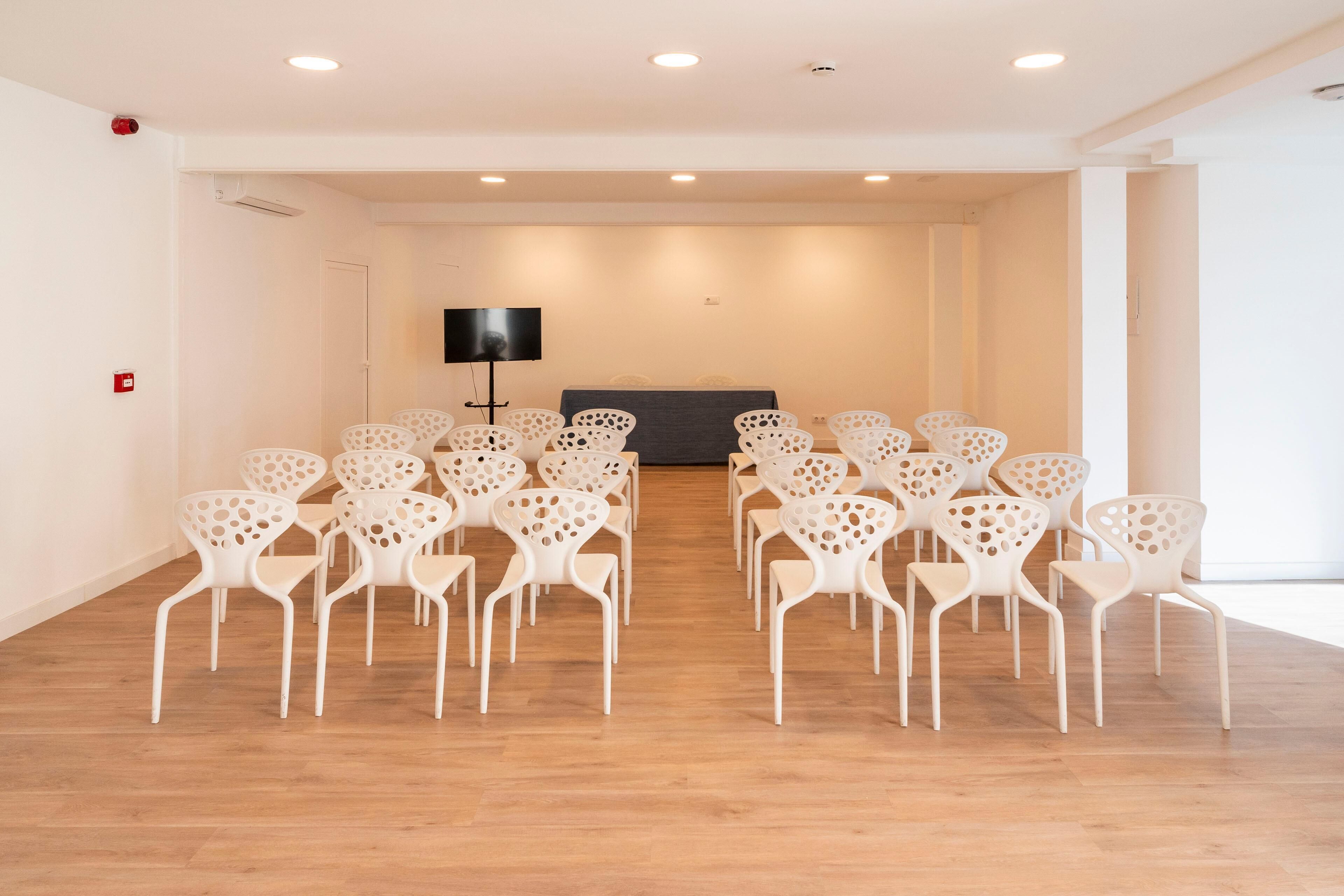 Een congreszaal met witte stoelen in rijen, een flatscreen televisie aan de muur en een houten vloer.