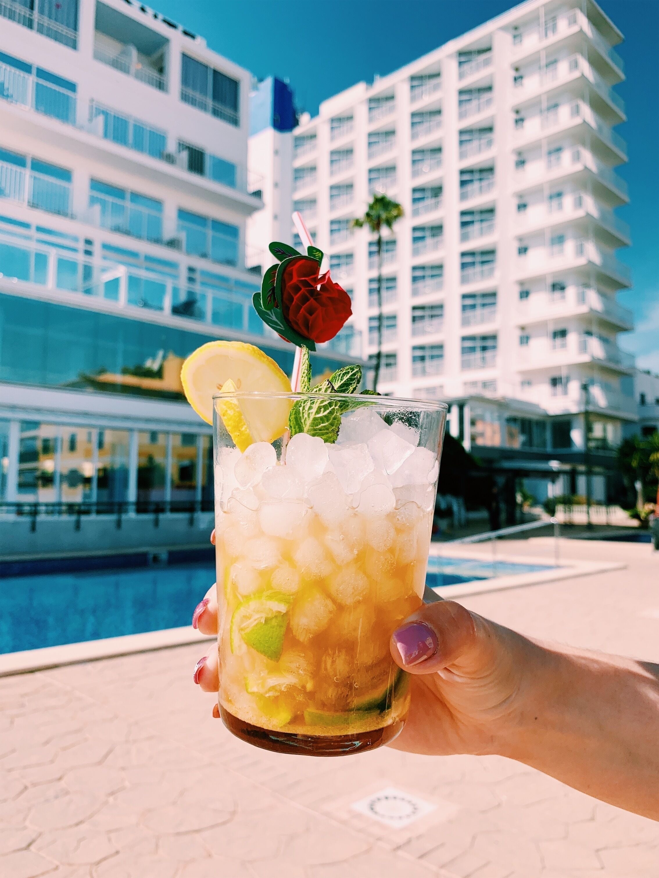 Een hand houdt een cocktail met ijs en garnering vast, met een modern gebouw en zwembad op de achtergrond.