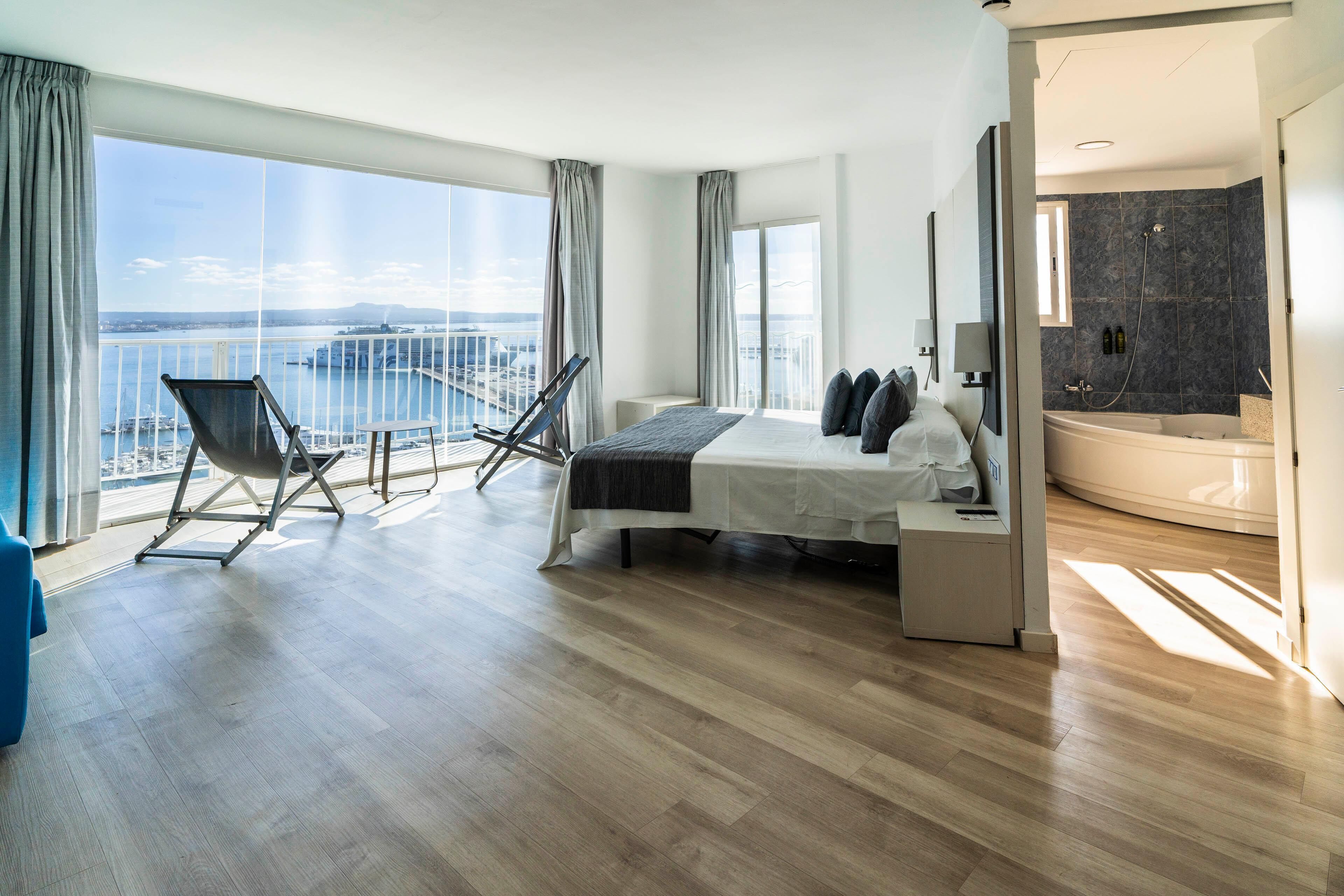 A modern hotel room with a large bed, a balcony with a view of the sea, a separate bathroom with a bathtub, and a seating area with chairs.