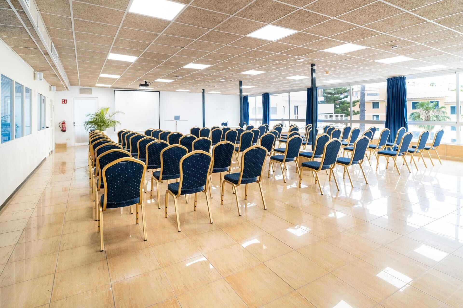 Une salle de conférence avec des rangées de chaises bleues, un projecteur et une grande fenêtre avec des rideaux.