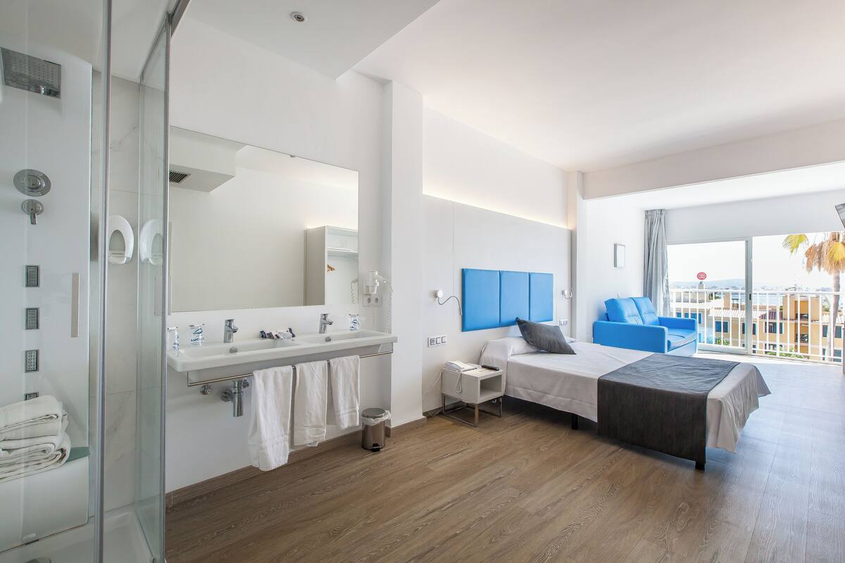 Une chambre d’hôtel moderne dotée d’une douche en verre, d’un grand lit avec une tête de lit grise, d’une table de chevet et offrant une vue sur la ville grâce à de grandes fenêtres.