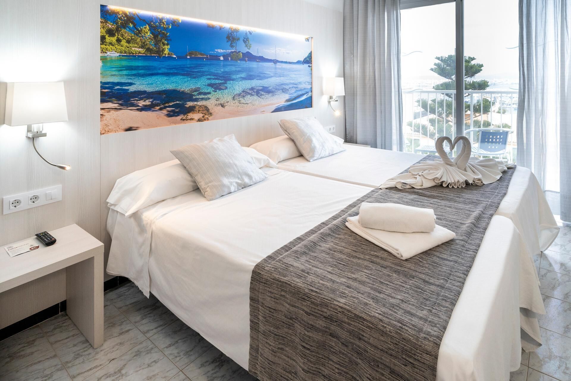 A hotel room with two beds, a large window with a view of the sea, a painting of a beach scene, and a bedside table with a lamp.