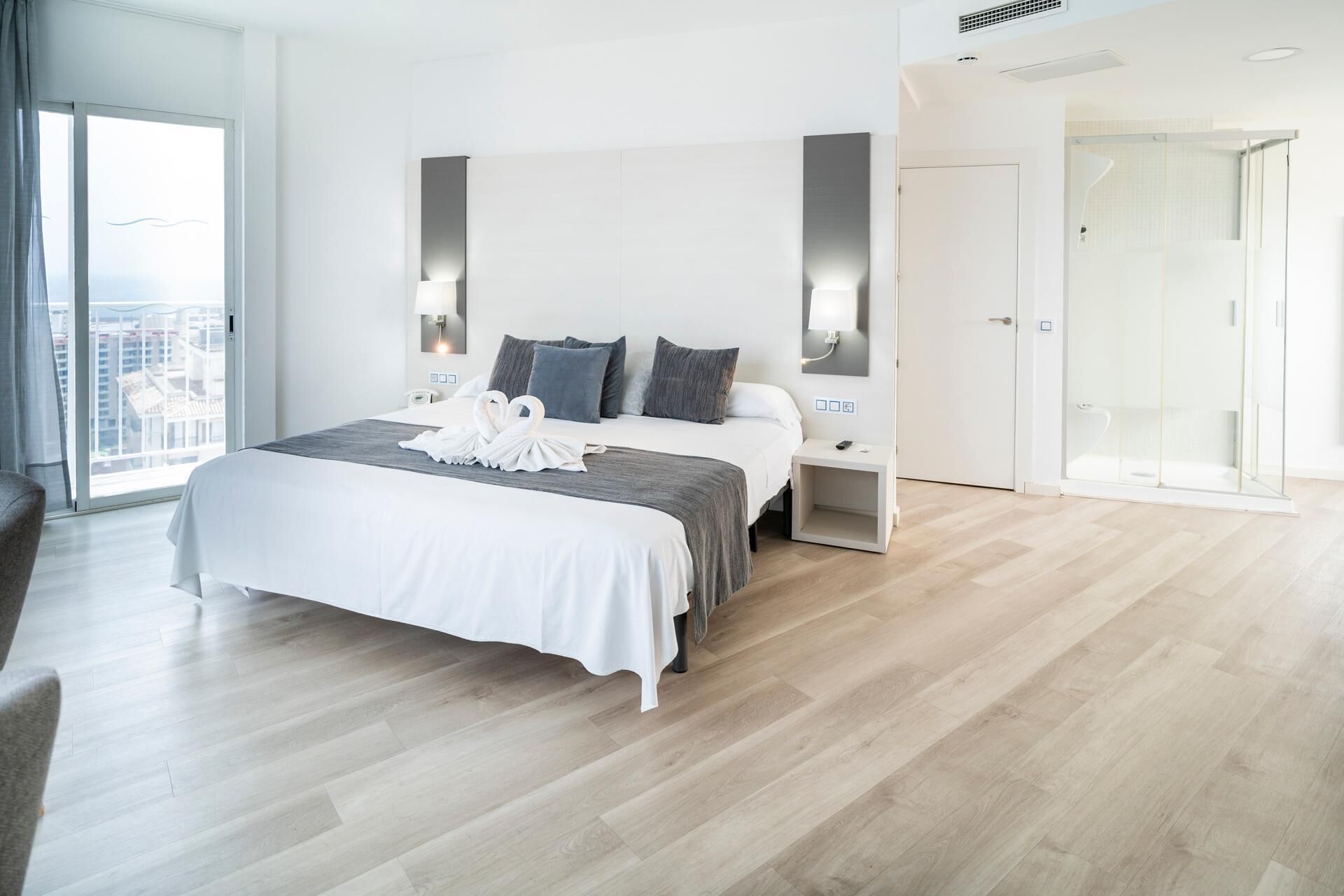 Habitación de hotel moderna con una cama grande, una mesita de noche y una cabina de ducha de vidrio, con vistas a la ciudad a través de un amplio ventanal.