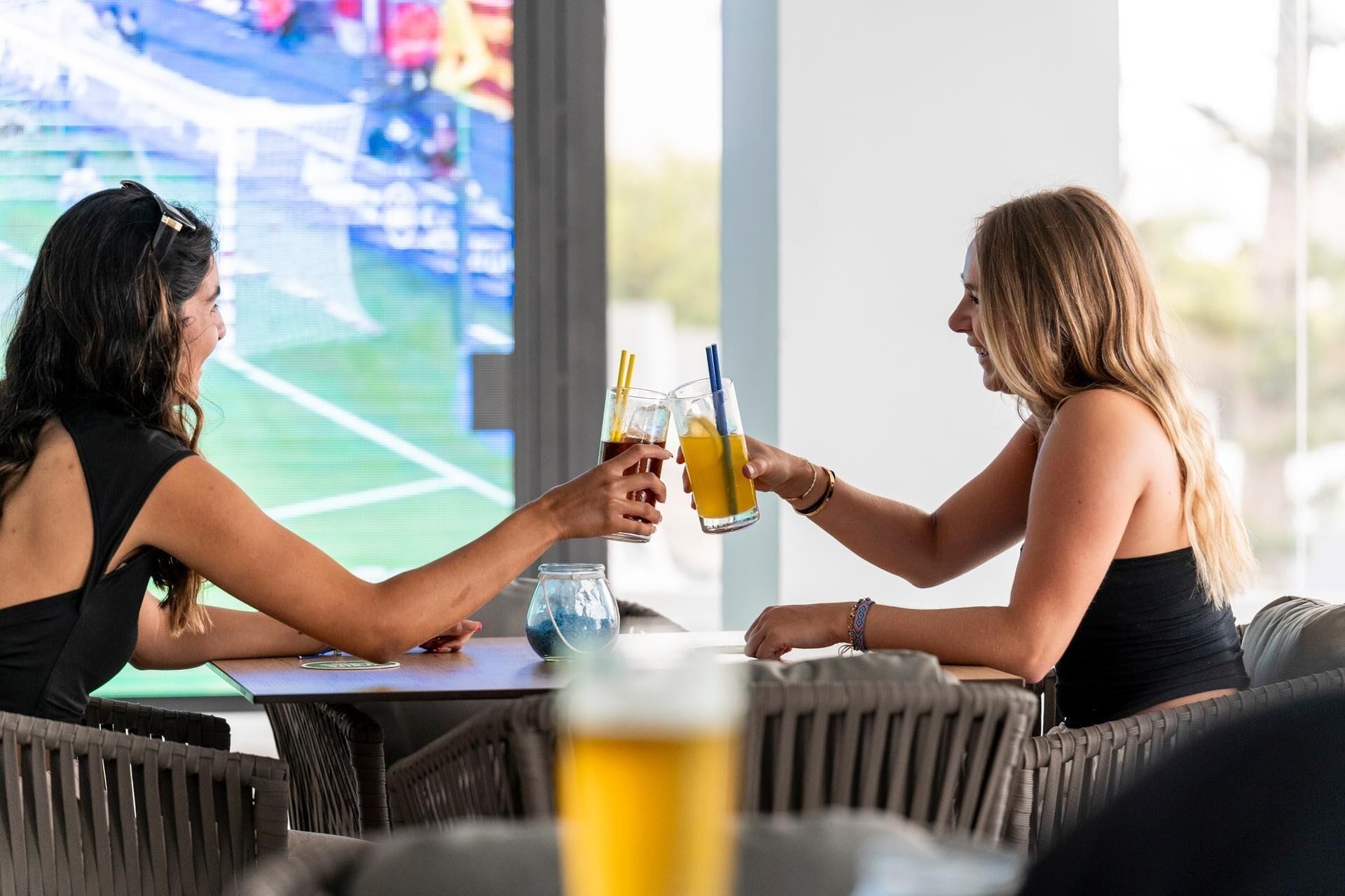 Dos personas brindando con bebidas en una mesa, con un vaso de agua y una botella de cerveza.