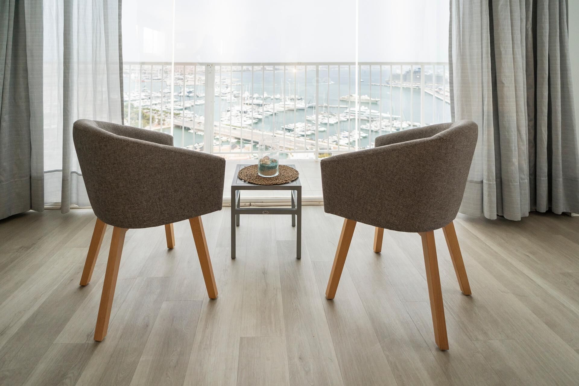Un salon moderne avec deux fauteuils gris, une petite table agrémentée d’un objet décoratif et une vue sur une marina, visible à travers de grandes fenêtres.