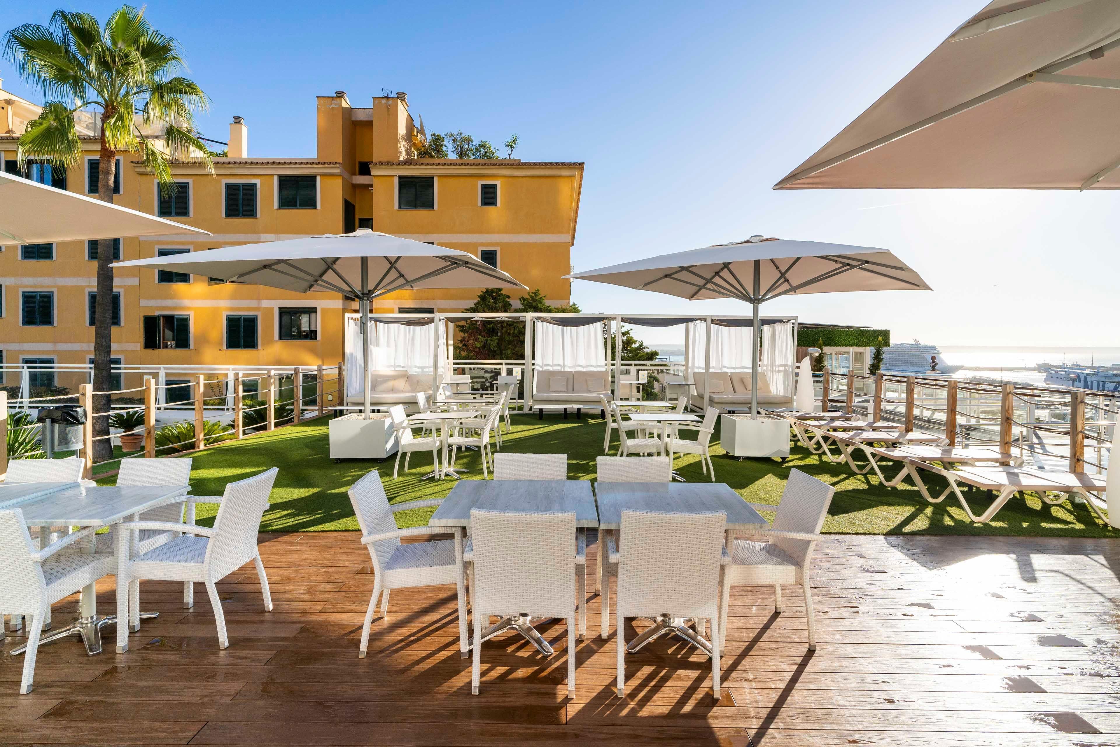 Eine Terrasse im Freien mit weißen Korbmöbeln und Sonnenschirmen, mit Blick auf eine grüne Rasenfläche und ein Gebäude mit Balkonen.