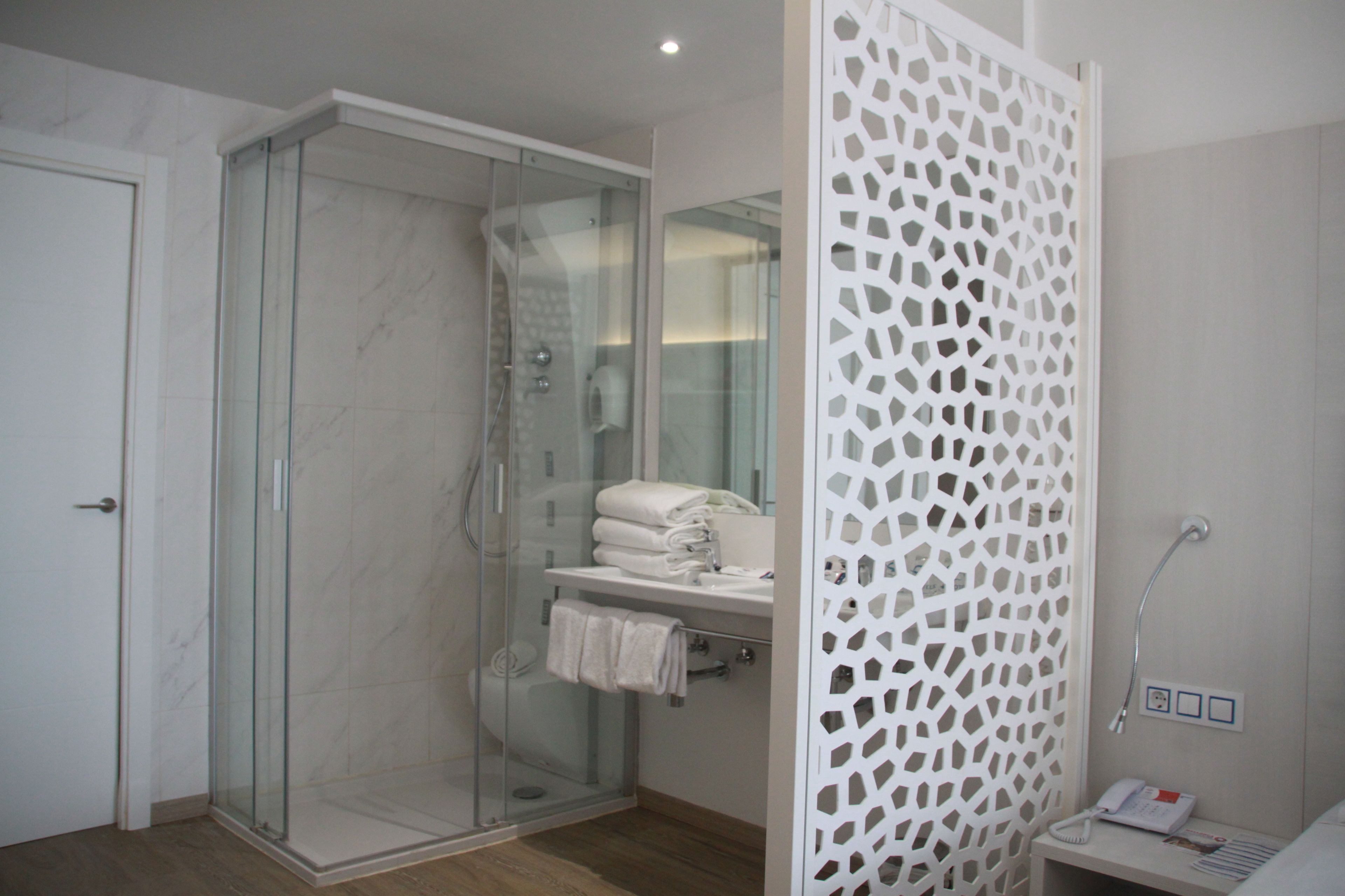 Un baño moderno con ducha envuelta en vidrio, un separador decorativo blanco y un pequeño tocador con toallero.