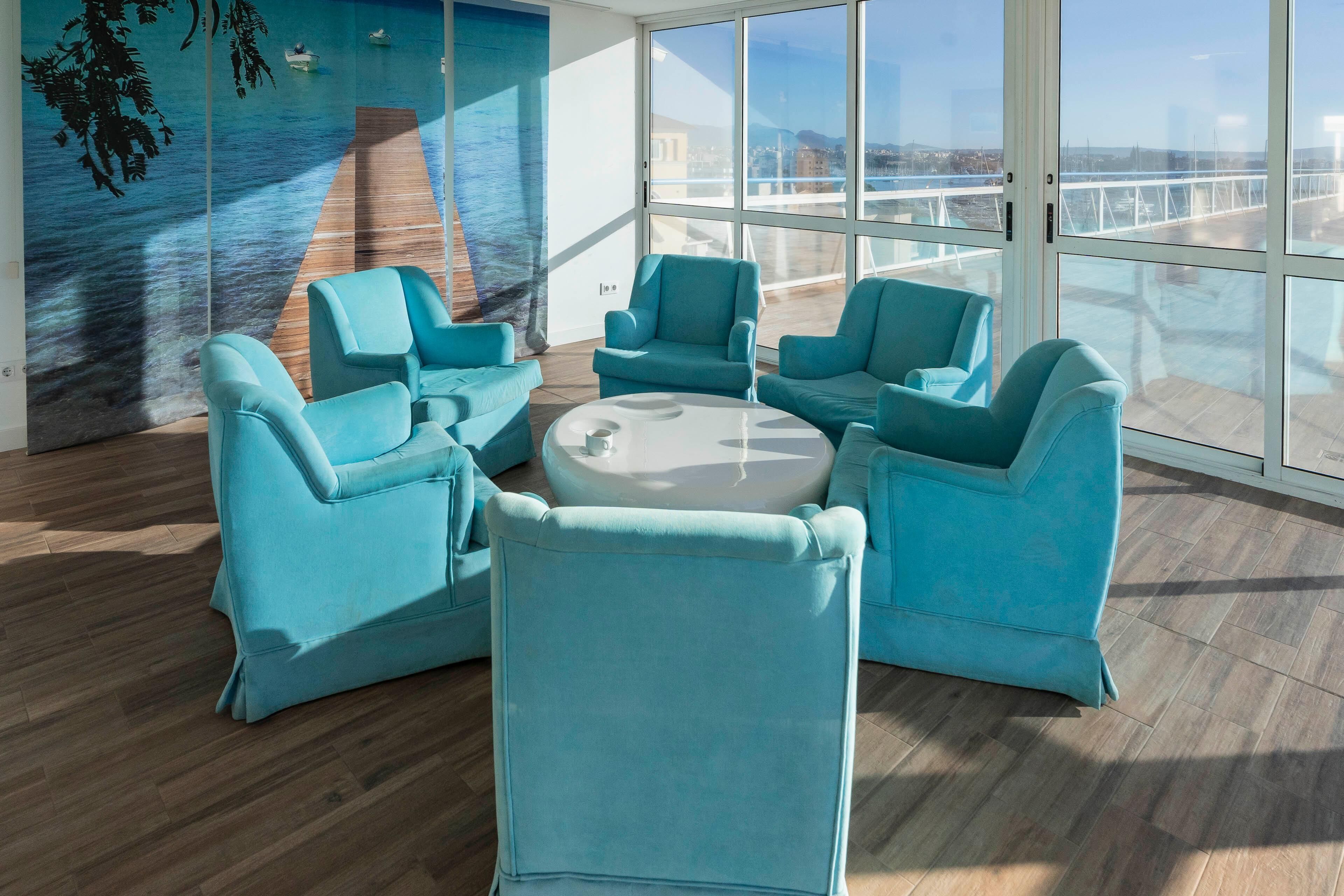 A modern lounge with teal chairs and a round table, large windows with a view of the sea and a bridge, and a mural of a ship on the wall.