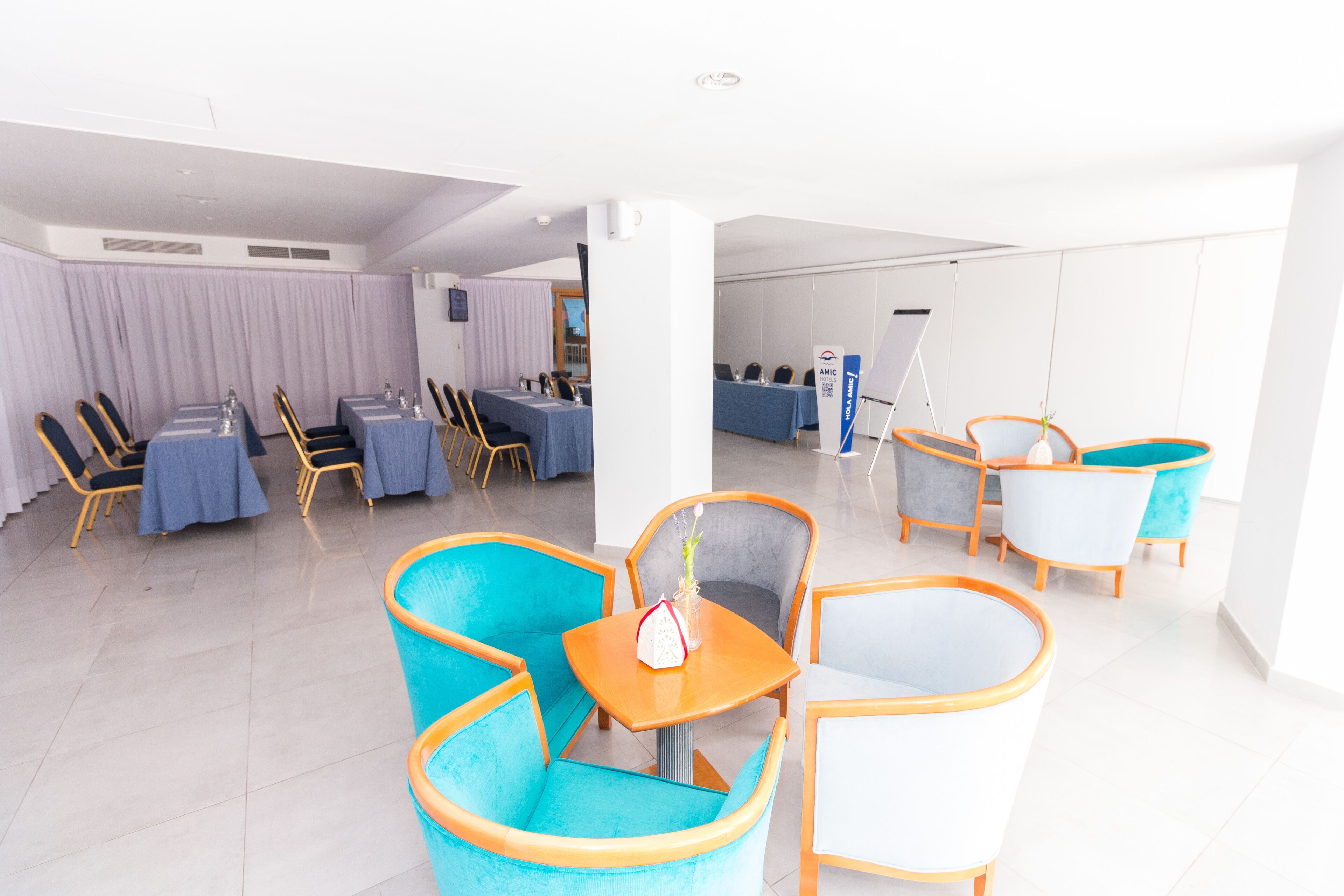 A modern hotel lobby with a seating area featuring teal and white chairs, a small table with a vase, and a reception desk with a blue tablecloth.
