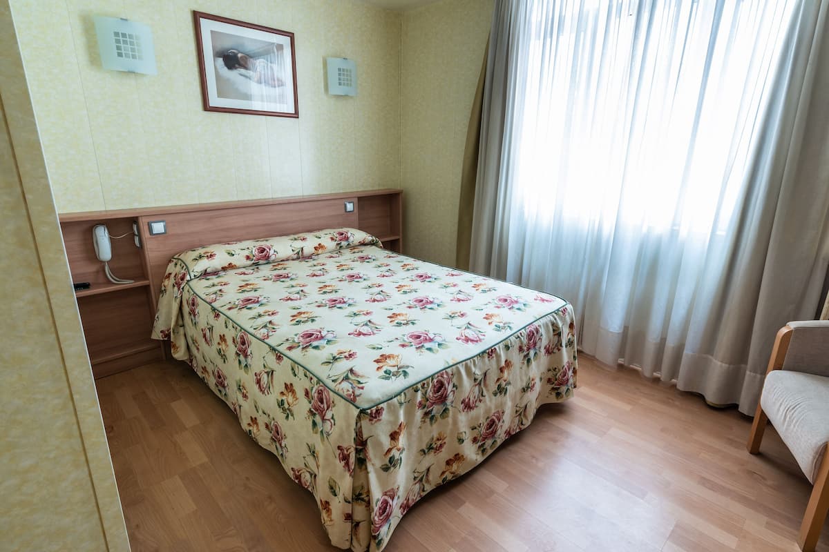 A hotel room with a floral bedspread, a wooden headboard, a bedside telephone, and a window with curtains.