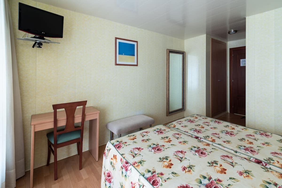 A hotel room with a wooden desk, a chair, a bed with a floral bedspread, a flat-screen TV mounted on the wall, a framed picture, and a mirror.
