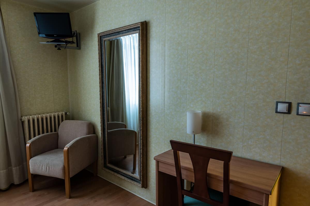 A hotel room with a large mirror, a wooden chair, a small table, a lamp, and a television mounted on the wall.
