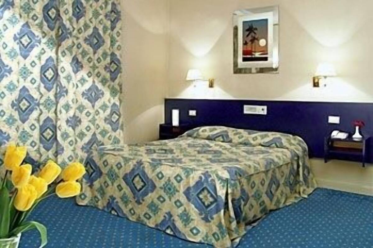 Habitación de hotel con una cama, una mesita de noche, una lámpara, un cuadro enmarcado en la pared y un jarrón con flores amarillas.