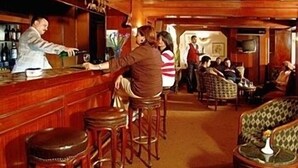 Una zona de bar con taburetes de madera y una barra. Se observan varias sillas y mesas al fondo.