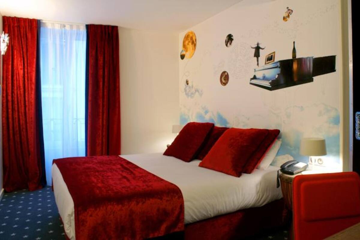A hotel room with a bed, red curtains, a desk with a chair, a wall clock, and a window with curtains.