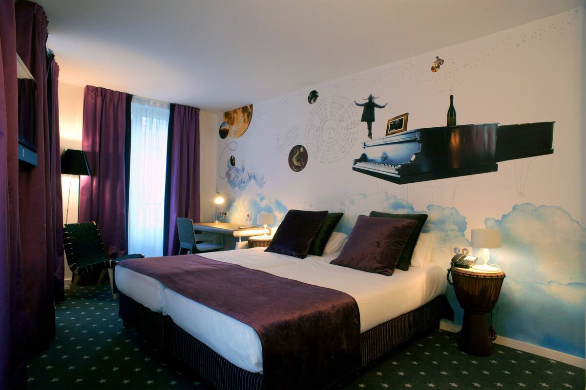 A hotel room with a large bed, a desk, a chair, a nightstand, and a wall with artistic decorations.