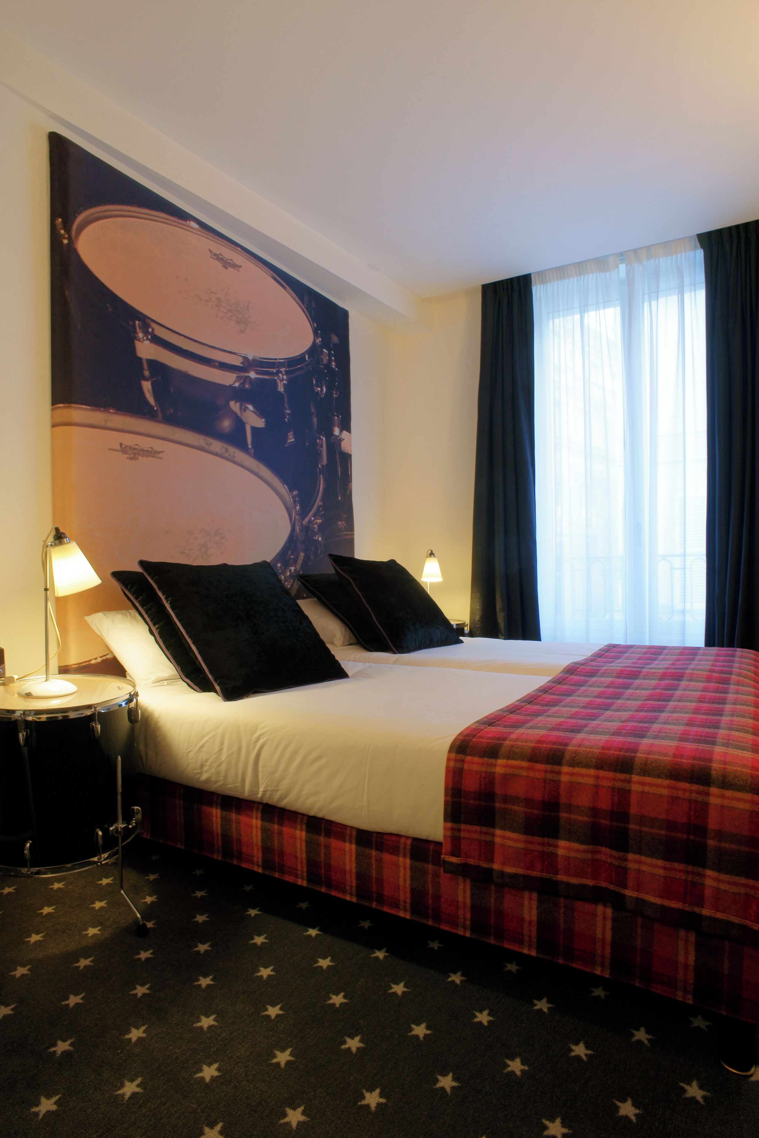 A hotel room with a bed, a drum set poster, a star-patterned carpet, and a window with curtains.