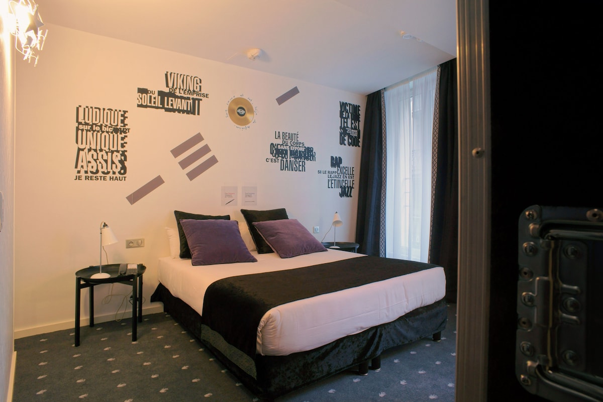Hotelkamer met een bed, nachtkastje, lamp en een muur met diverse tekst en symbolen.