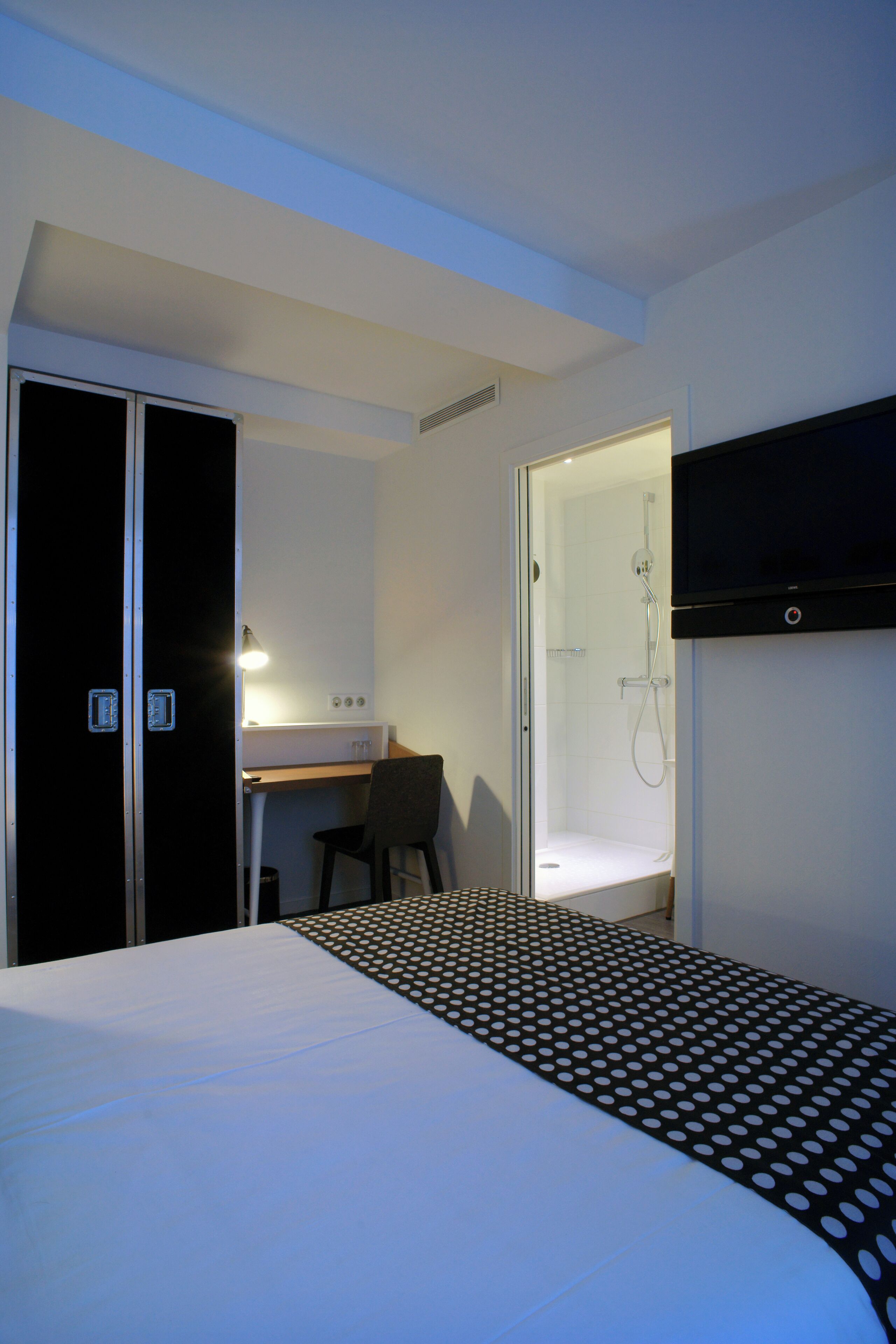 A hotel room with a bed, a desk, a chair, and a shower area.