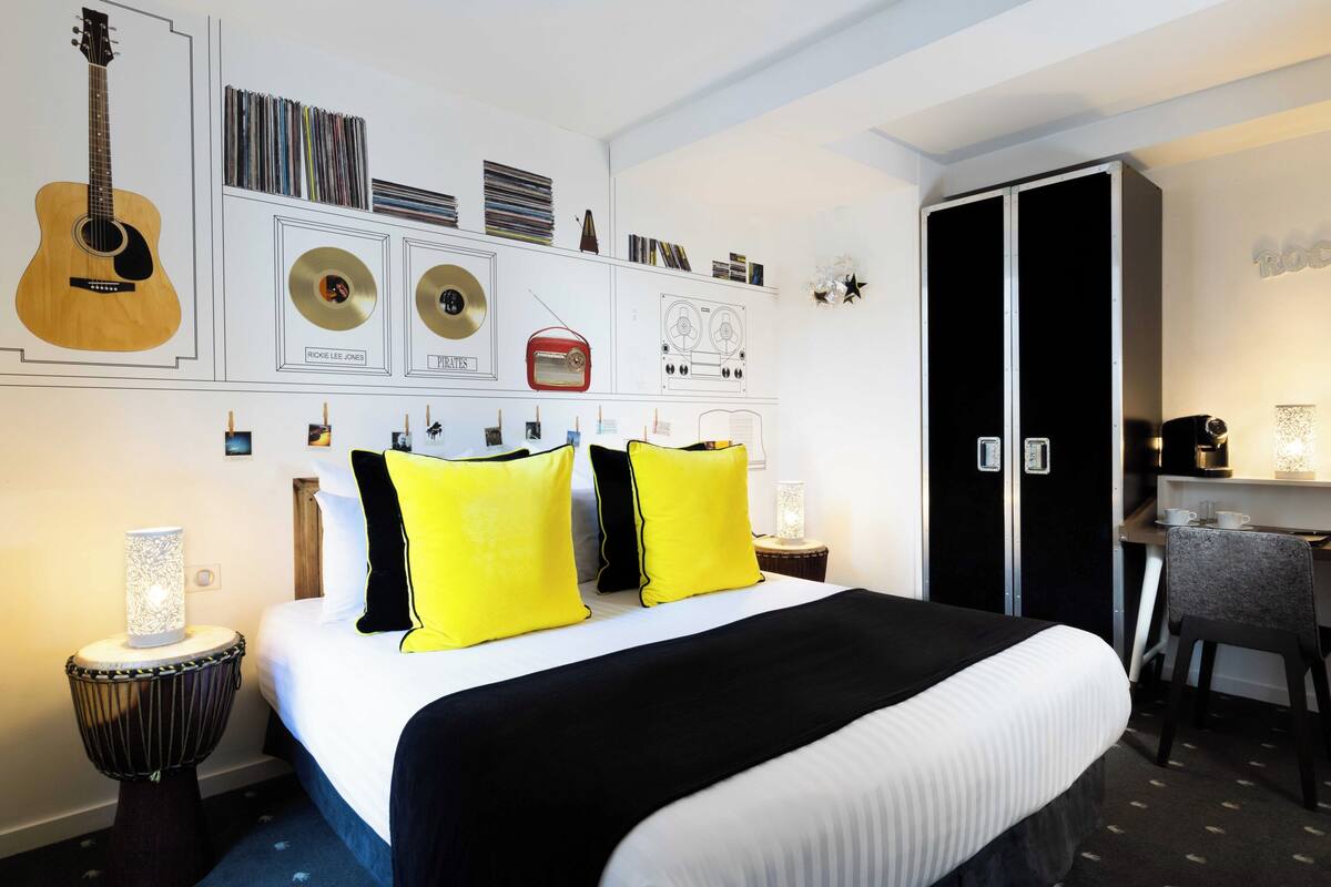 A hotel room with a bed, a desk, a wardrobe, and wall decorations including a guitar, records, and a radio.