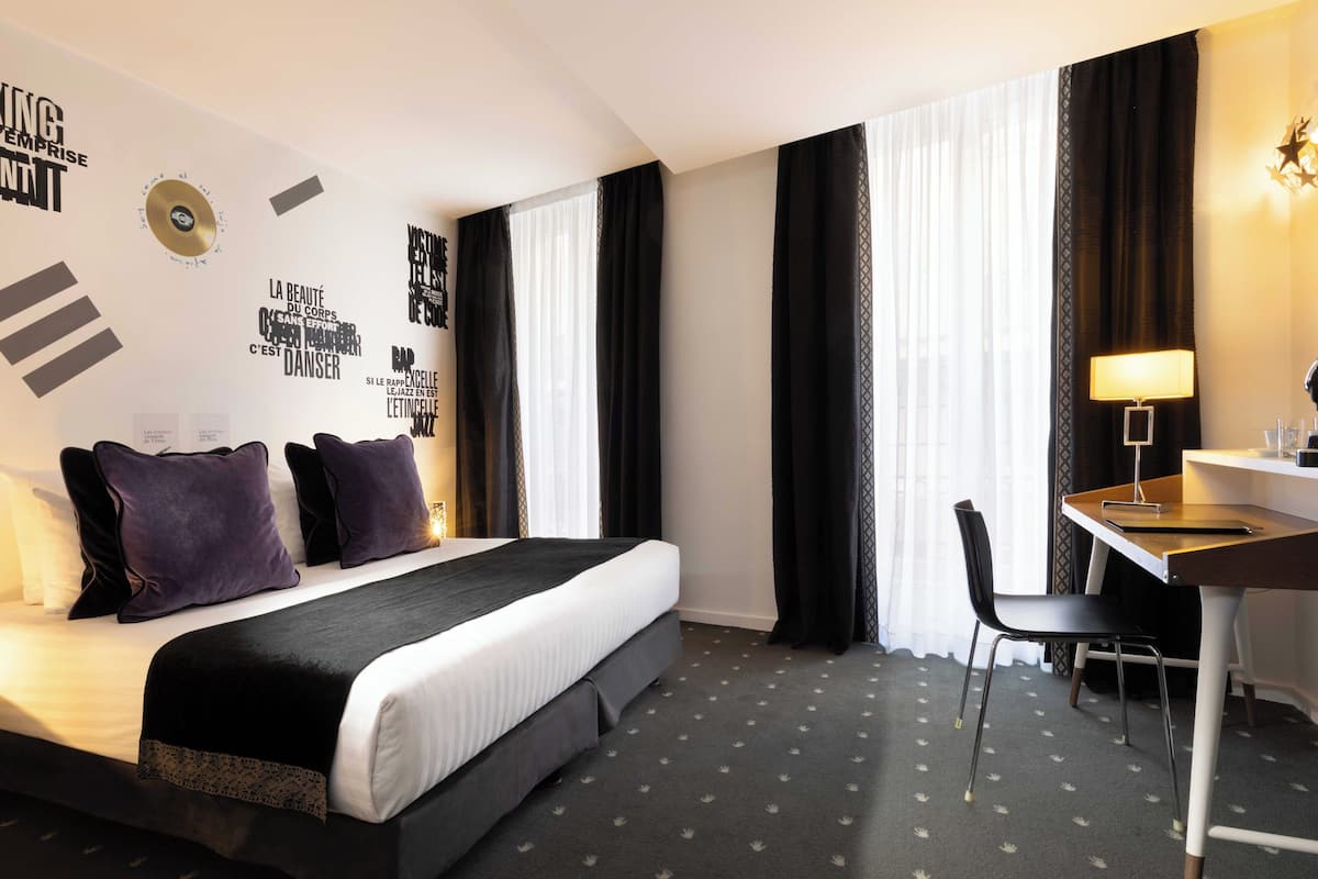 A hotel room with a bed, a desk, and a chair. There are curtains on the windows, and a wall with text and a star decoration.