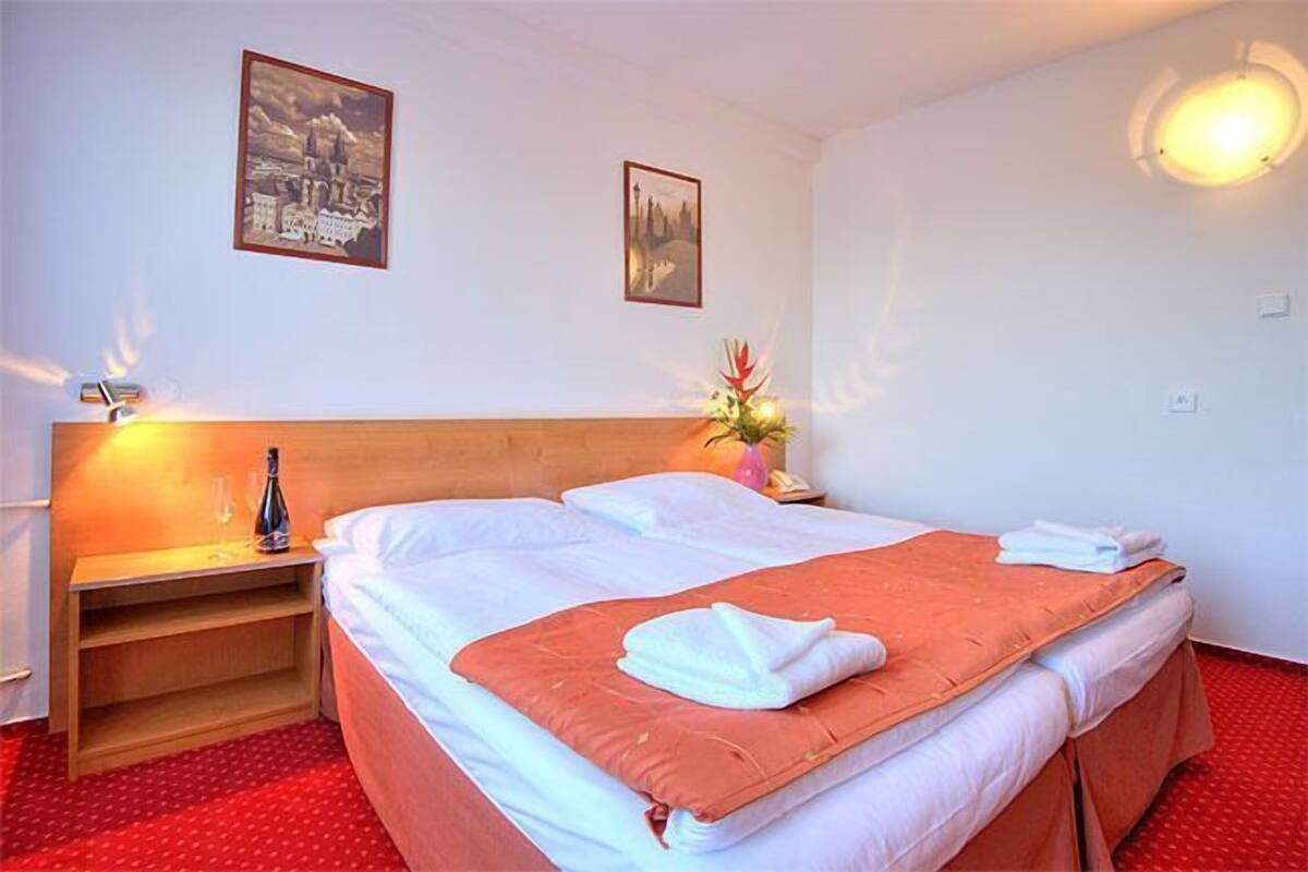A hotel room with a bed, a nightstand, two framed pictures on the wall, and a red carpet.